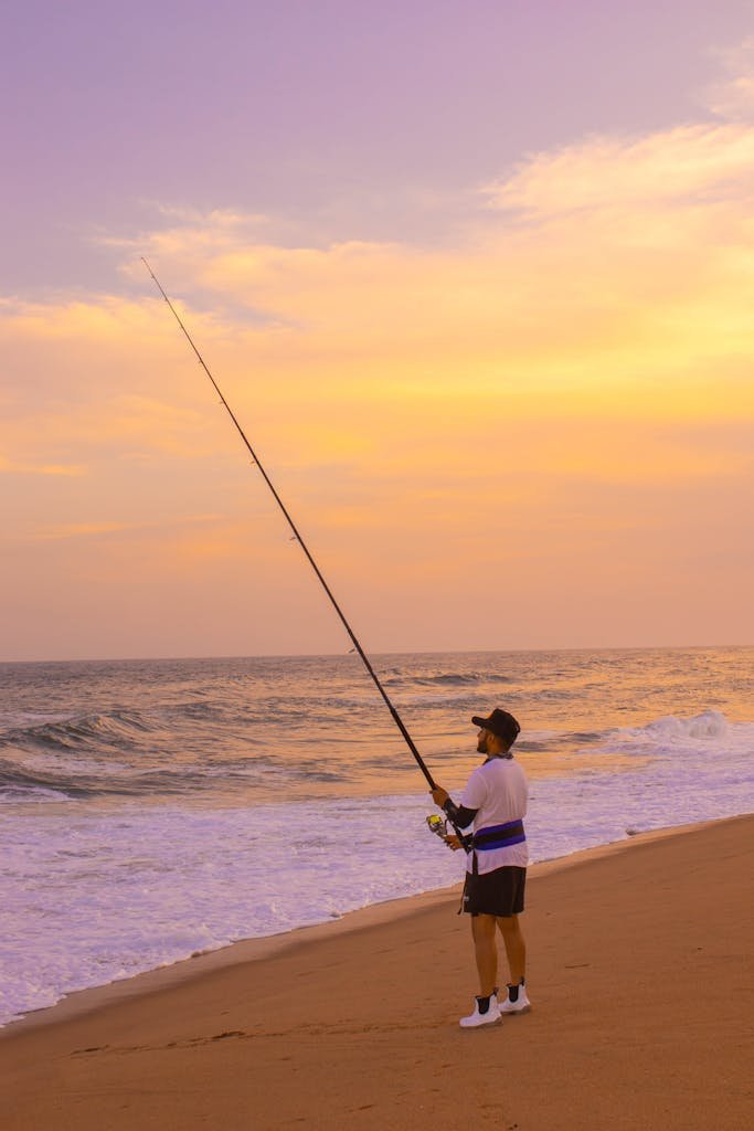 A fisherman casts his line on a sandy beach during a vibrant sunset.