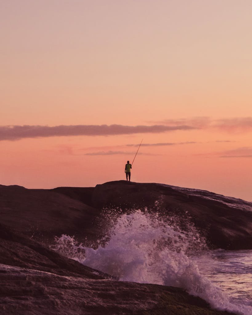 A fisherman silhouetted against a vibrant sunrise on a rocky ocean cliff.