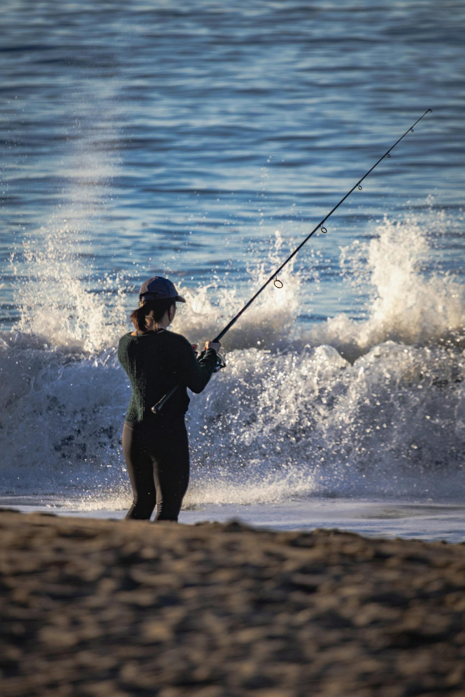 A person fishes at the ocean's edge as waves crash around them, creating a dynamic scene.