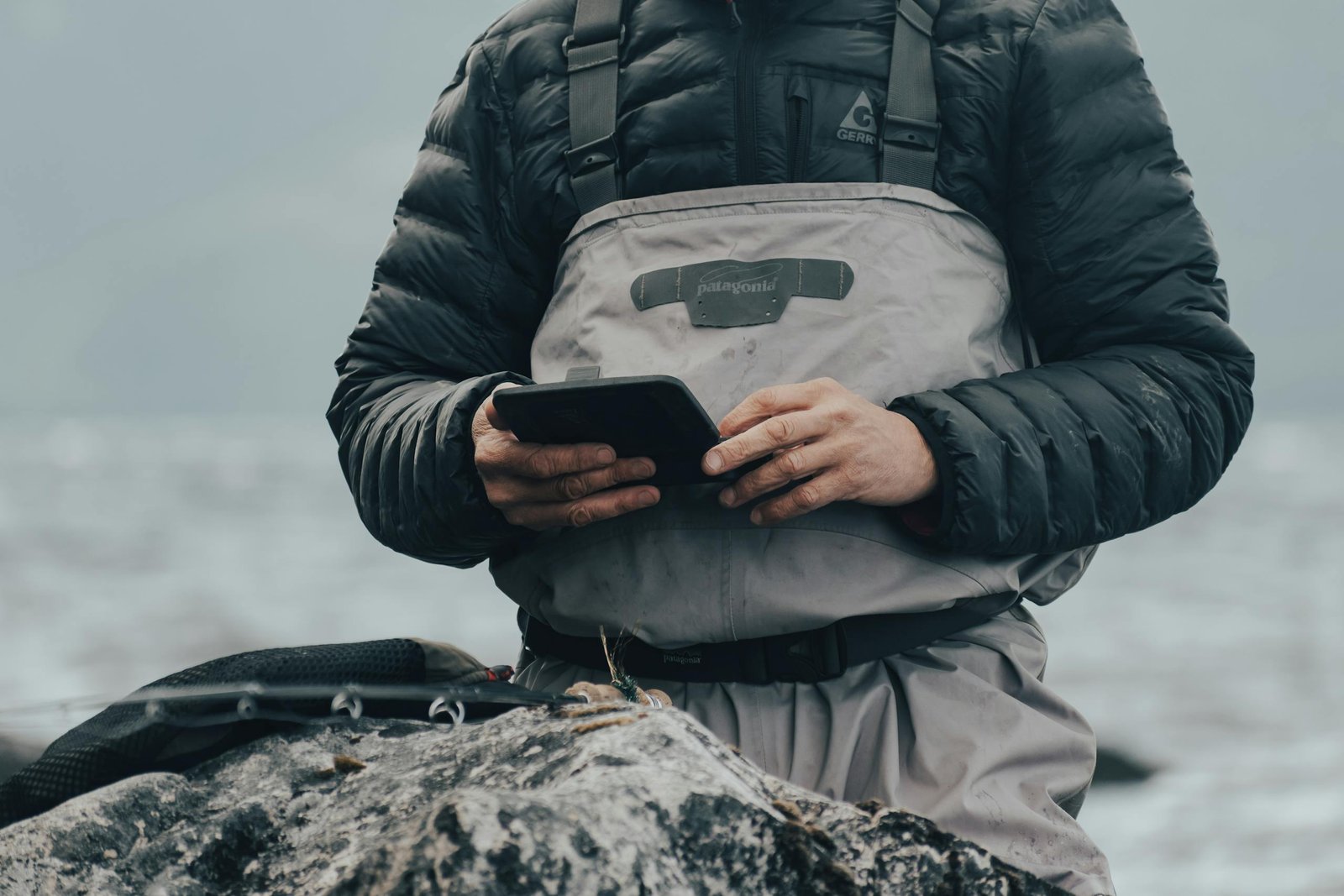 Angler in a jacket using fishing equipment by the water's edge.